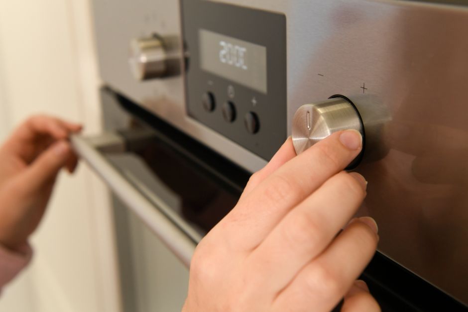 Close-up image of a person's hand adjusting the temperature dial on a stainless steel electric oven in a domestic kitchen. The oven features a digital display showing a temperature setting of 250°C, 