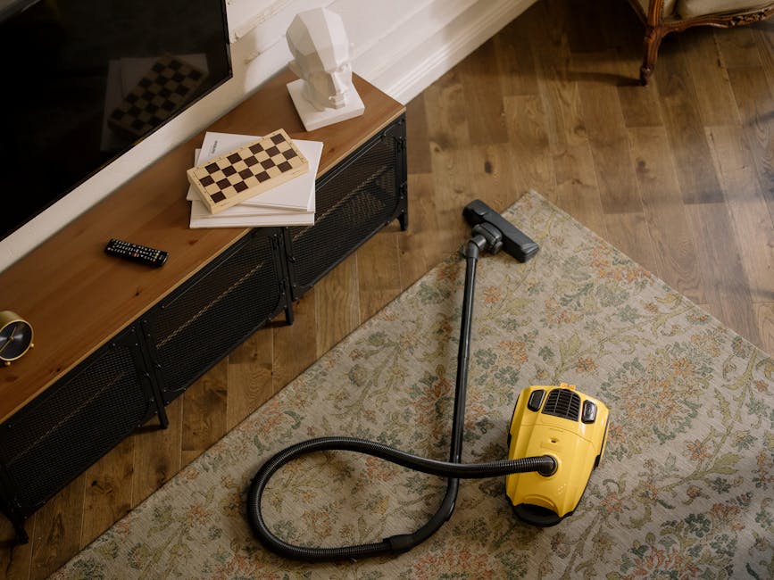 A woman using a yellow vacuum cleaner equipped with a hose attachment to deep clean a patterned area rug in a living room. The rug features an intricate floral design in soft shades of beige, light bl